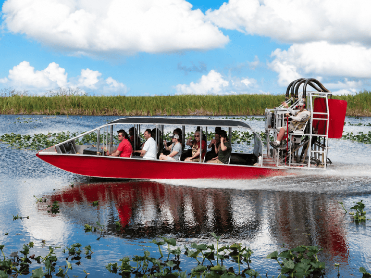 Red airboat with passengers gliding through a marsh under a blue sky with clouds.